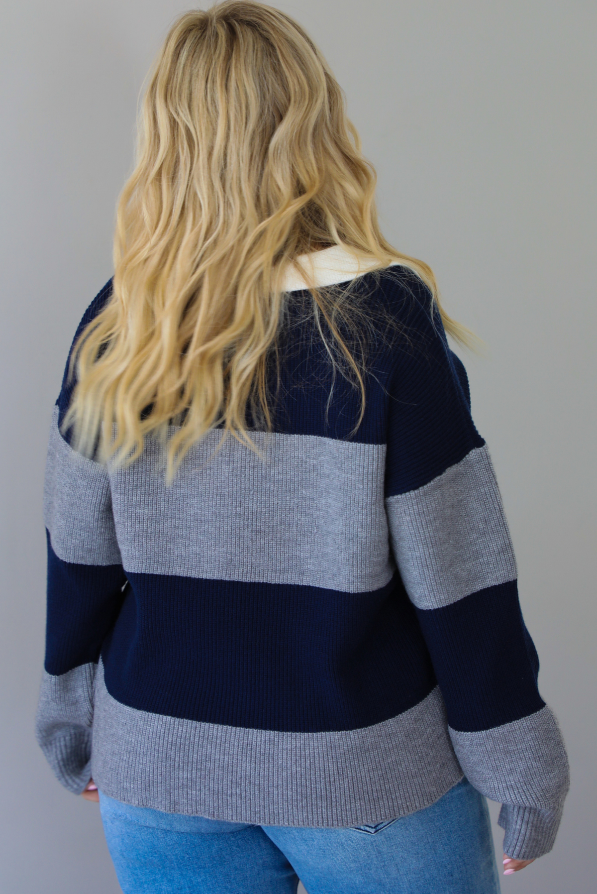A woman shown from behind wearing a navy and charcoal boyfriend style pullover top with a white collared detail, with distressed denim, standing indoors against a white wall.