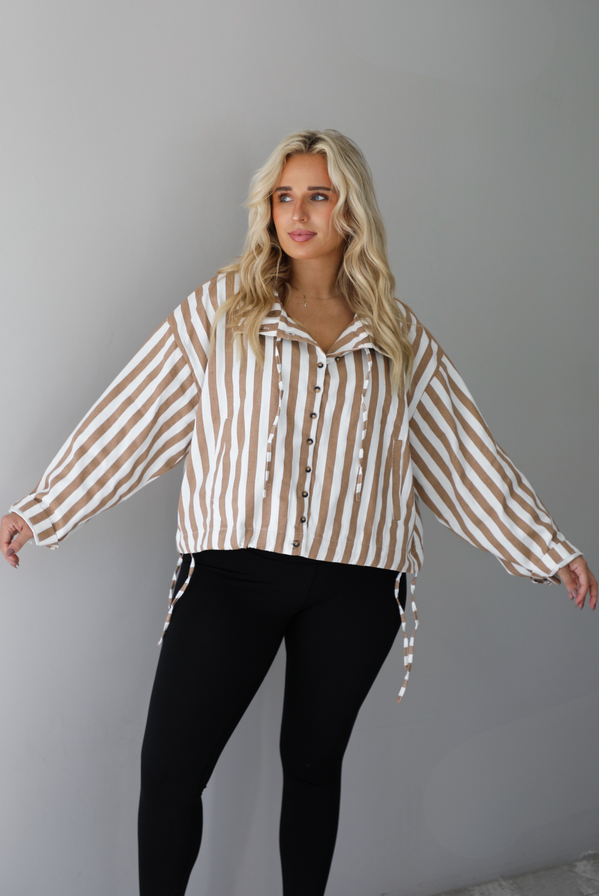 A woman wearing a tan and white striped collared long sleeved top with functional button closures along the front, worn with black leggings, standing indoors against a white wall. The tops drawstring details are un-cinched to show the full length. 