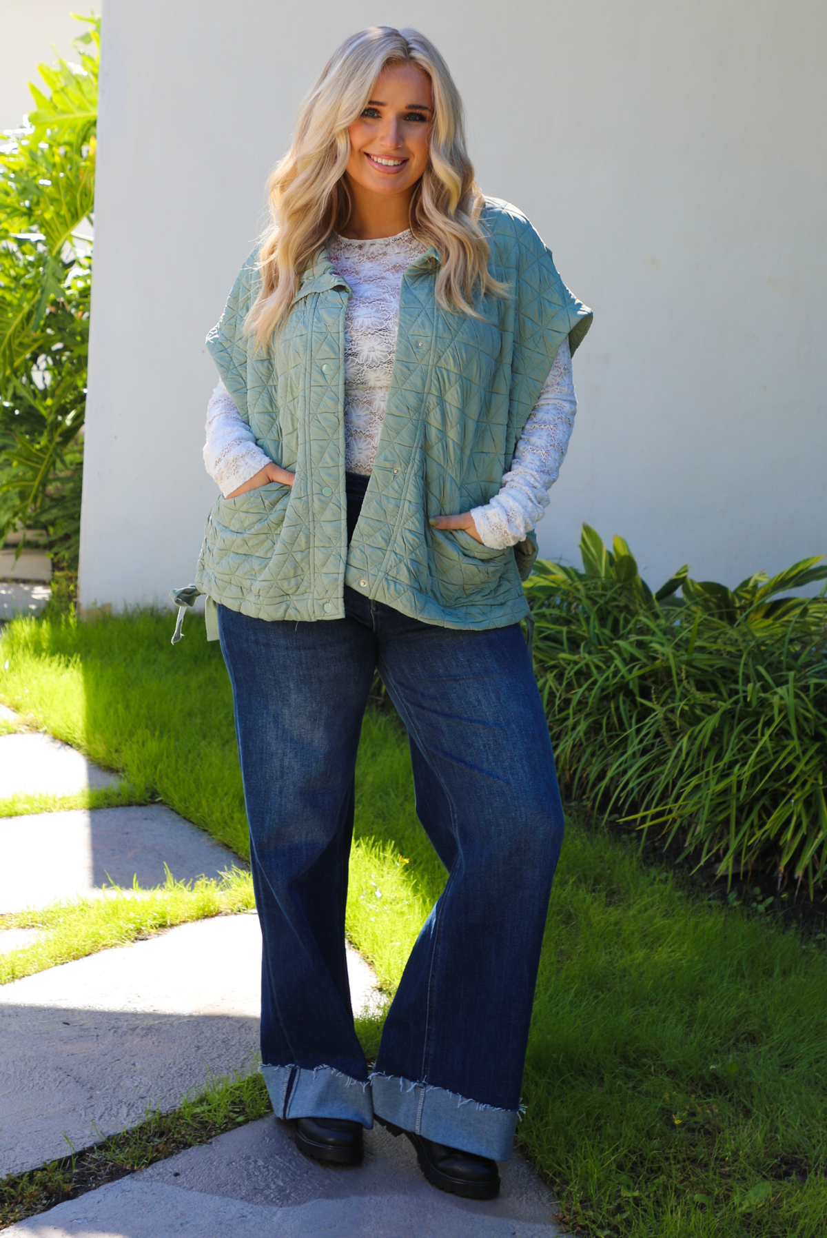 Woman model wearing a sage colored oversized vest paired with a white lace long sleeved top and dark denim outdoors.  The vest has functional pockets on either side, functional button closures in the front, and an oversized fit. 