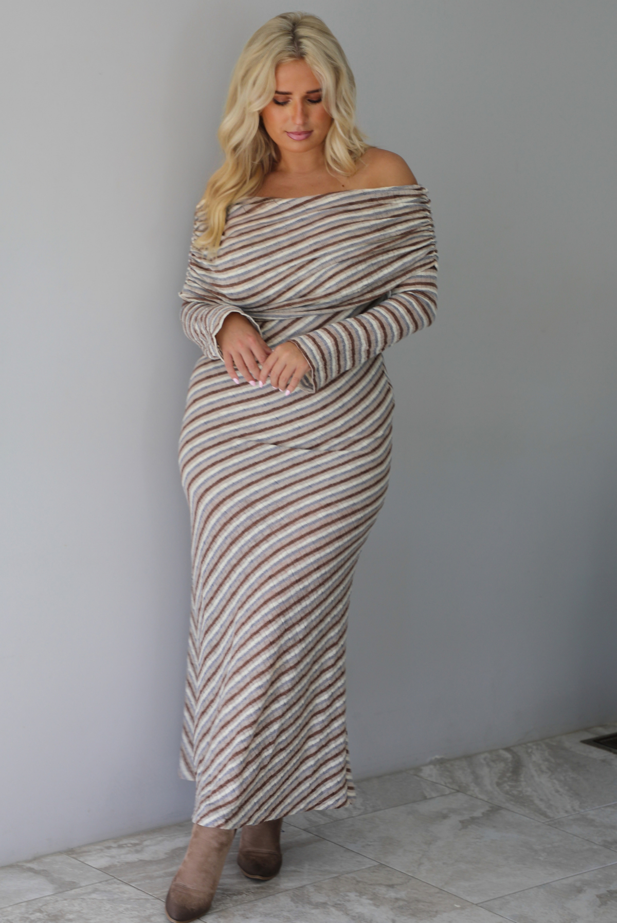 Woman wearing a grey, brown, muted blue, and off white striped fitted maxi dress that has a folded down design at the shoulders and bodice and long sleeves, standing indoors against a white wall with white marble flooring. 