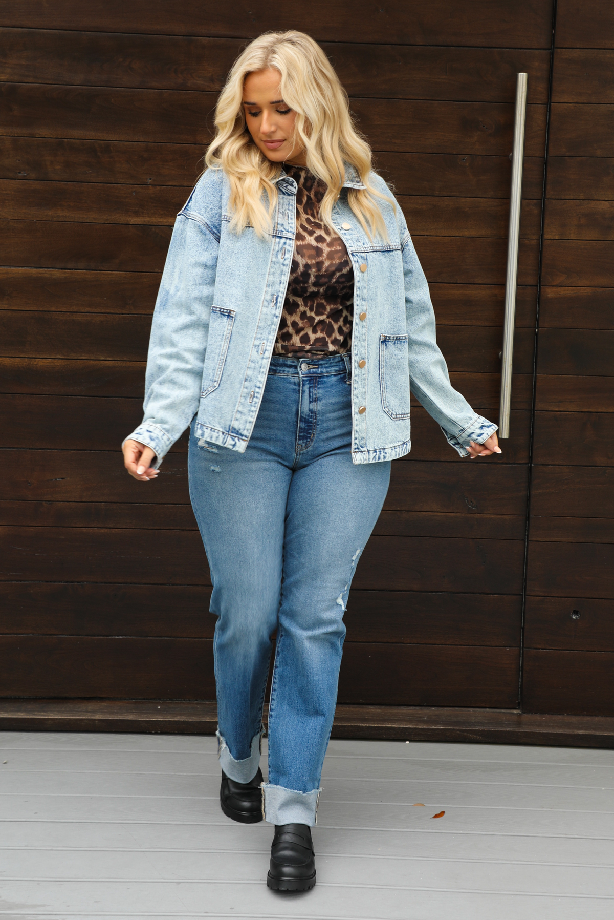 Woman model wearing an oversized, acid washed, light denim jacket, paired with a sheer mesh leopard print top, medium wash denim jeans, and black shoes. The photo is taken outdoors against a dark brown door. The model is taking a step forward while looking to her right.