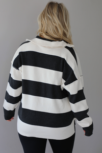 Woman wearing a charcoal and white striped quarter-zip sweater with a collared neckline, raw seam details, and a relaxed fit. The photo shows the back of the sweater. It's paired with a fitted black jumpsuit. The photo is taken indoors against a white wall with white marble flooring.