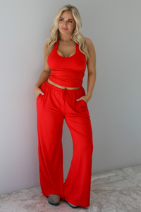Woman wearing a red two piece set featuring a  tank top and slouchy pants. The photo is taken indoors against a white wall with white marble flooring. 