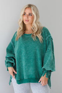Woman wearing a green colored oversized sweatshirt with exposed seam details and a high-low design with heather grey casual pants. The photo is taken indoors against a white wall.