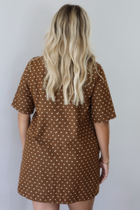 A woman with long blonde wavy hair is shown from behind wearing a brown short-sleeve collared dress with a cream polka dot pattern, indoors against a white wall. 