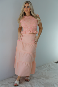 Woman wearing a pink and pale yellow gingham print two piece set featuring a smocked top and high waisted, maxi length skirt. The photo is taken indoors against a white wall with white marble flooring. 