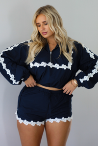 A woman modeling a matching navy blue two-piece set with white floral lace trim, consisting of a cropped zip-up jacket and shorts, standing indoors against a white wall.