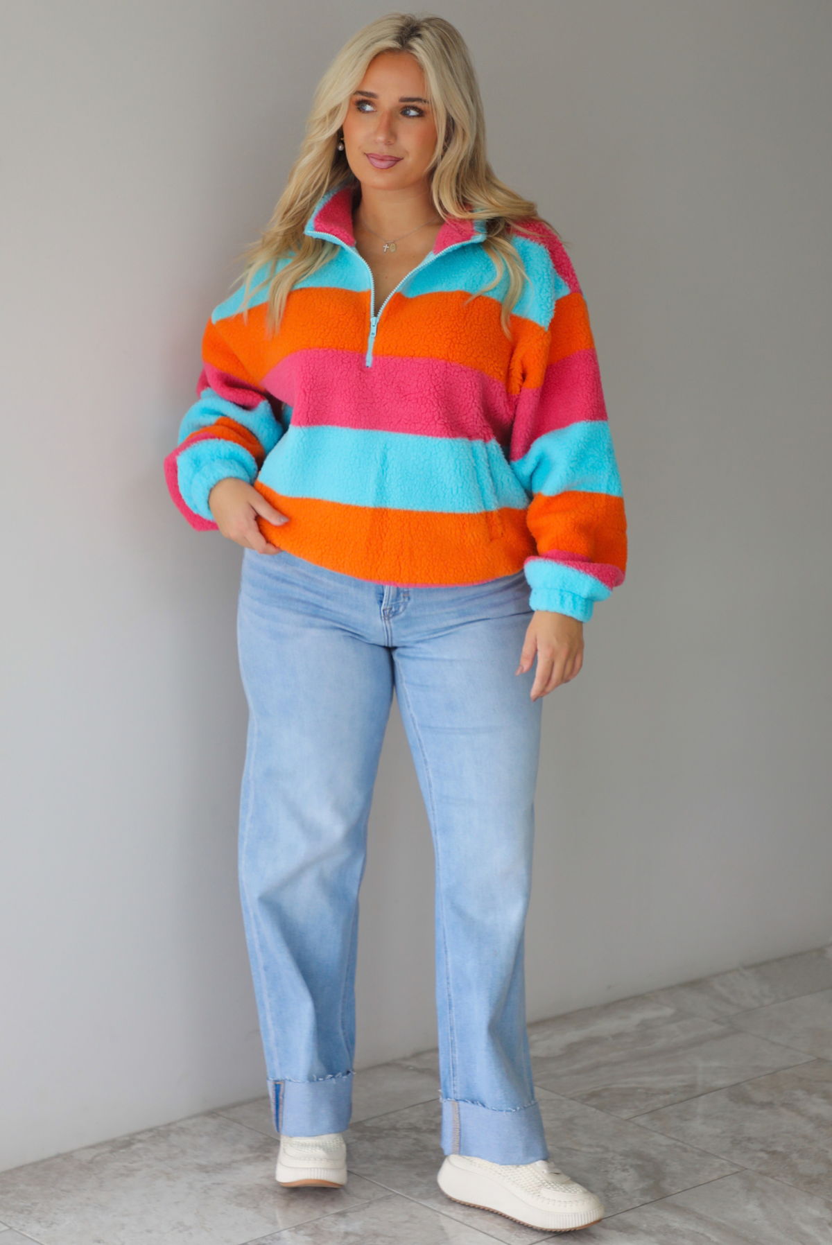 A woman wearing a turquoise, pink, and orange striped sherpa quarter-zip sweater with light wash denim jeans, standing indoors against a white wall with white marble flooring. 