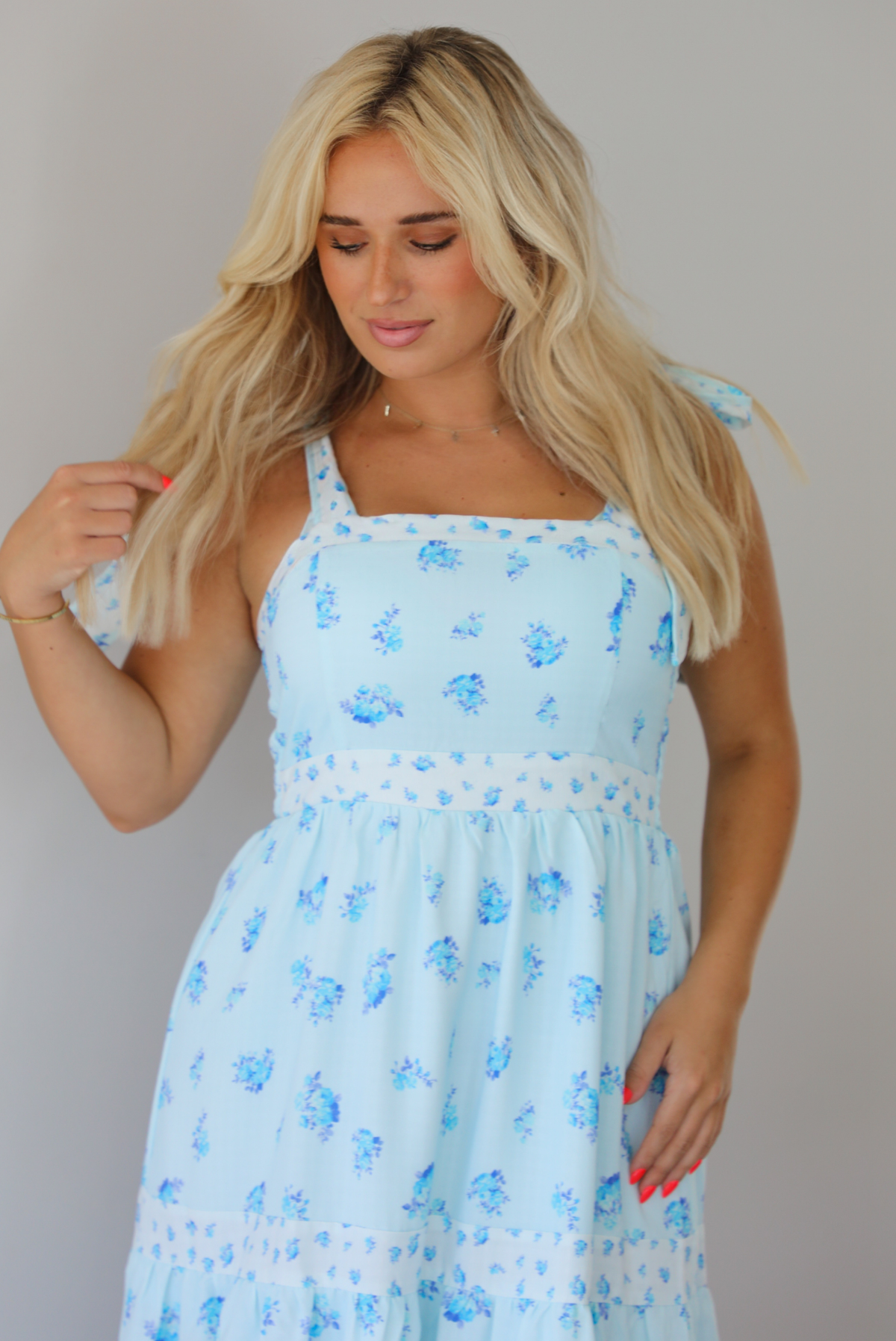 Woman wearing a light blue floral maxi dress indoors against a white wall. The photo shows a closeup of the bodice of the dress, which has a cinched waistline detail. 