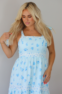 Woman wearing a light blue floral maxi dress indoors against a white wall. The photo shows a closeup of the bodice of the dress, which has a cinched waistline detail. 