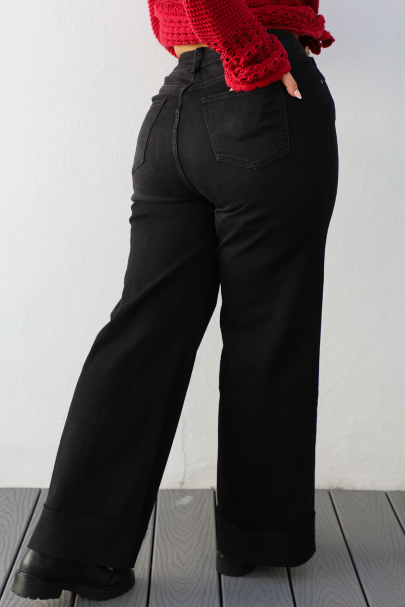 Back of a woman model wearing a black denim jeans outdoors against a white wall. The jeans are high waisted with button and zipper closures in front, have functional front and back pockets, belt loops around, and a cuffed hem. This model is wearing a Large.