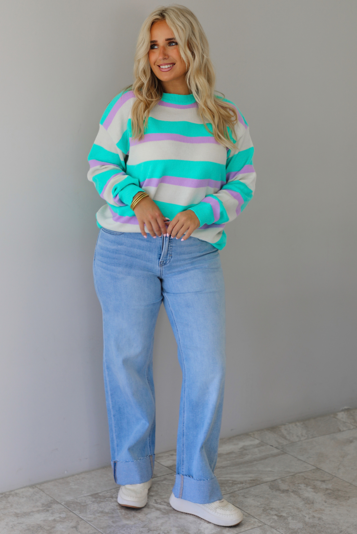 A woman wearing a turquoise, lavender, and cream striped knit sweater, paired with light wash denim jeans, standing indoors against a white wall with white marble flooring. 
