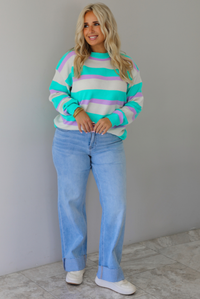 A woman wearing a turquoise, lavender, and cream striped knit sweater, paired with light wash denim jeans, standing indoors against a white wall with white marble flooring. 
