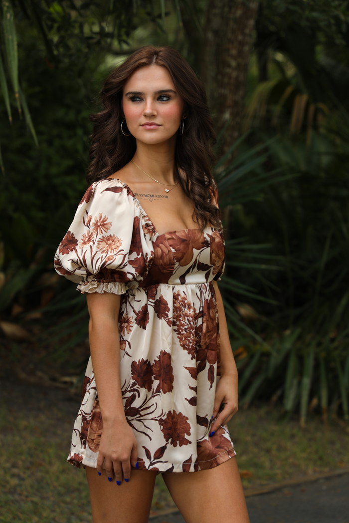 Woman model wearing an ivory and brown floral print satin romper that has puffed, short sleeves, a fitted bodice, and flowy shorts. The romper is paired with brown cowboy boots. The photo is taken outdoors on a paved road with greenery in the back. The photo shows a close up of the side of the romper and the puffed sleeves that cinch on the bicep with elastic. 