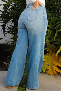 Woman model wearing a light wash denim that are high waisted, have functional front and back pockets, functional button closures in the front, distressed details, and a flared, almost bell-bottom style hem. The photo is taken outdoors against a white wall with greenery in the back. The photo showcases the back of the denim.