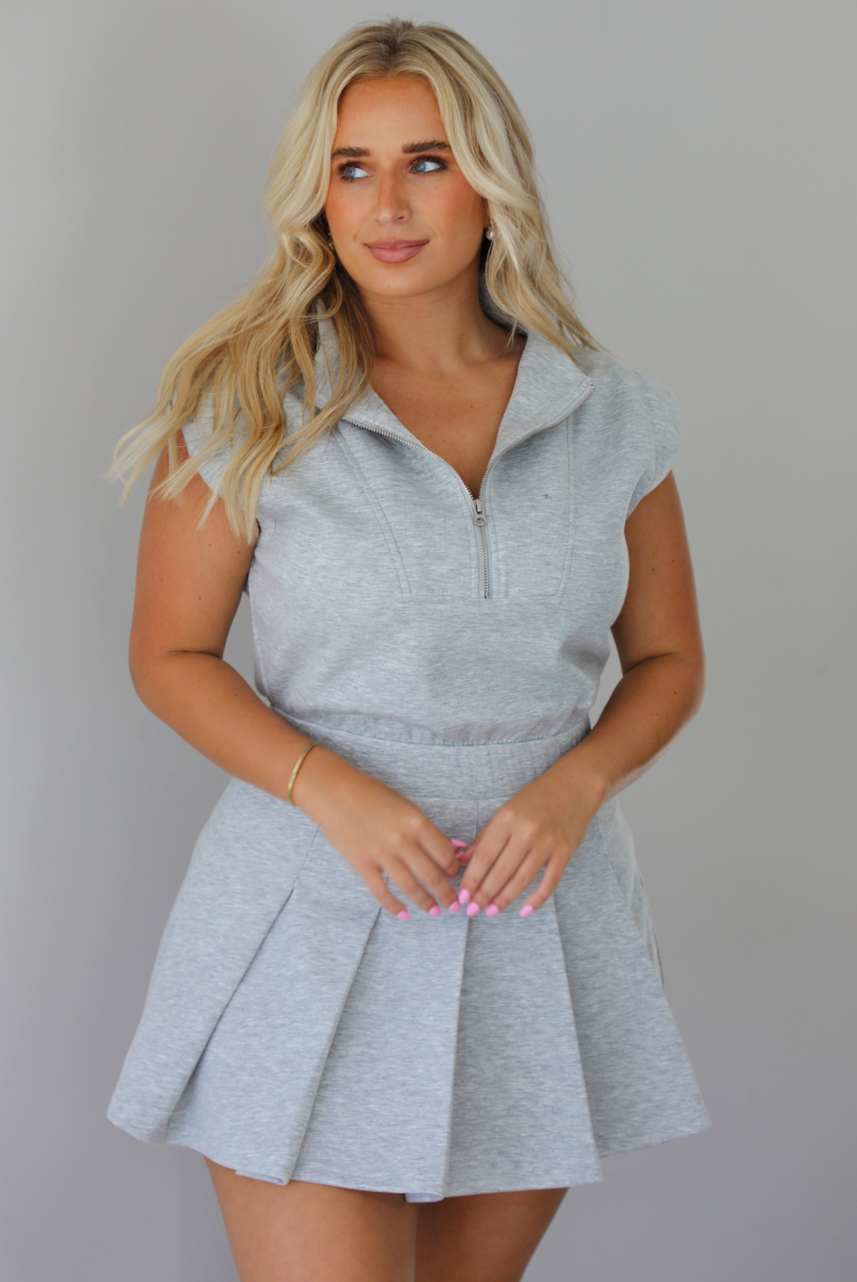 A woman modeling a light gray heathered short-sleeve collared dress with a pleated mini skirt,  indoors against a white wall.