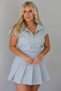 A woman modeling a light gray heathered short-sleeve collared dress with a pleated mini skirt,  indoors against a white wall.