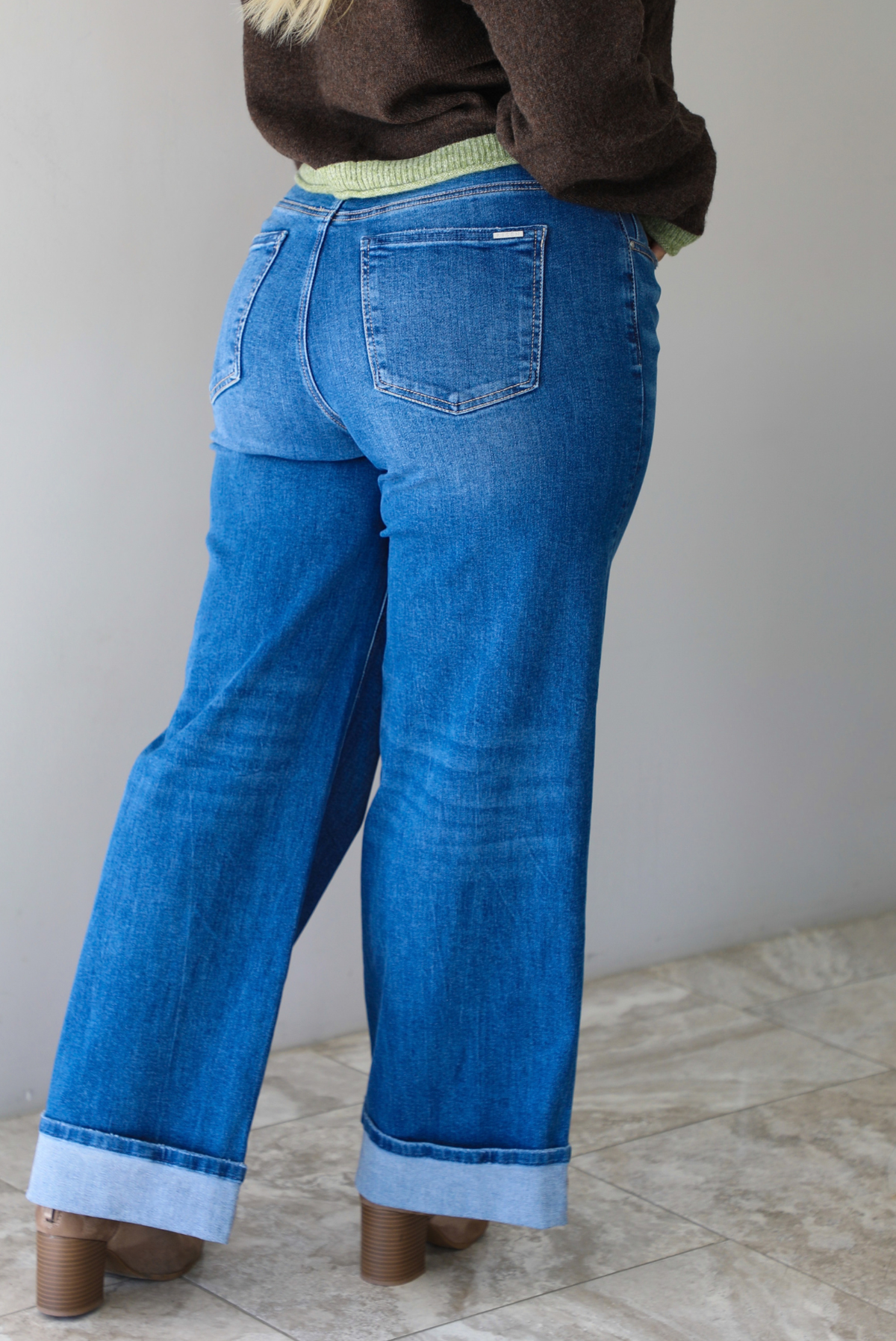 Woman shown from behind wearing a pair of dark wash denim jeans, standing indoors against a white wall with white marble flooring. 