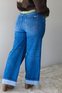 Woman shown from behind wearing a pair of dark wash denim jeans, standing indoors against a white wall with white marble flooring. 