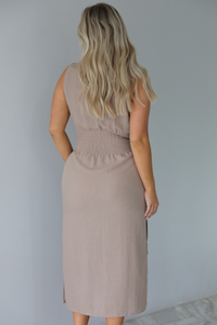 Woman wearing a tan, high neckline, midi dress that has a tie detail at the waistline and split hem details on either side. The photo is taken indoors against a white wall and shows the back of the dress, which has smocked material at the waistline in the back. 