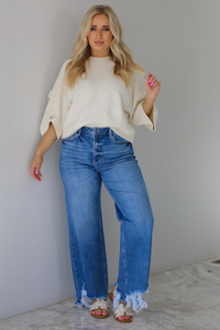 Woman wearing a cream colored quarter sleeve sweater that has ribbed details at the neckline, on the sleeves, and on the hem, with non-functional button closures on the sleeves. The sweater has a relaxed fit and is paired with a medium wash, high waisted, distressed hem denim with sand colored sandals. The photo is taken indoors against a white wall with white marble flooring. 