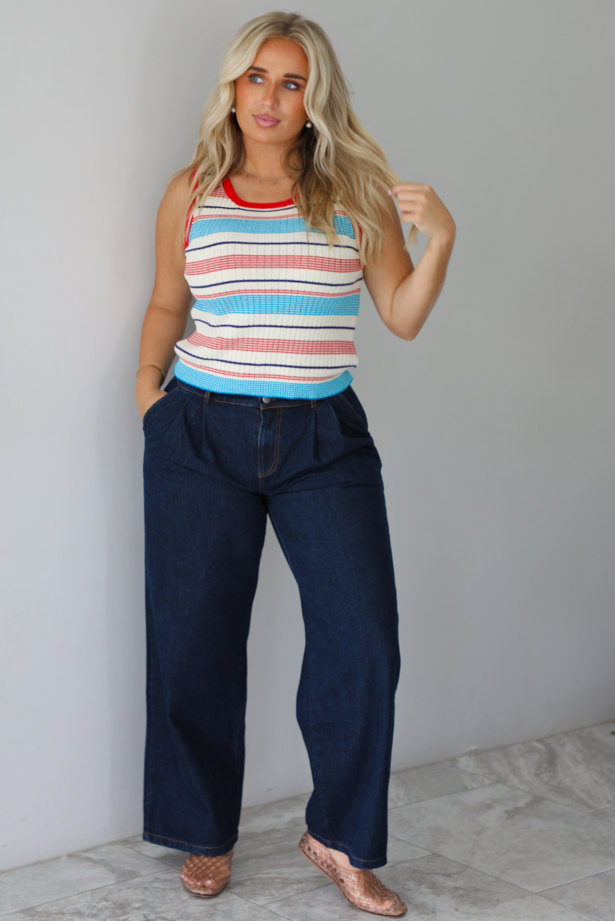 A woman modeling a sleeveless ribbed knit top with horizontal stripes in red, light blue, navy, and white, paired with dark wide-leg jeans and metallic rose gold flats, standing indoors against a white wall with white marble flooring. 