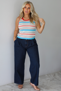 A woman modeling a sleeveless ribbed knit top with horizontal stripes in red, light blue, navy, and white, paired with dark wide-leg jeans and metallic rose gold flats, standing indoors against a white wall with white marble flooring. 