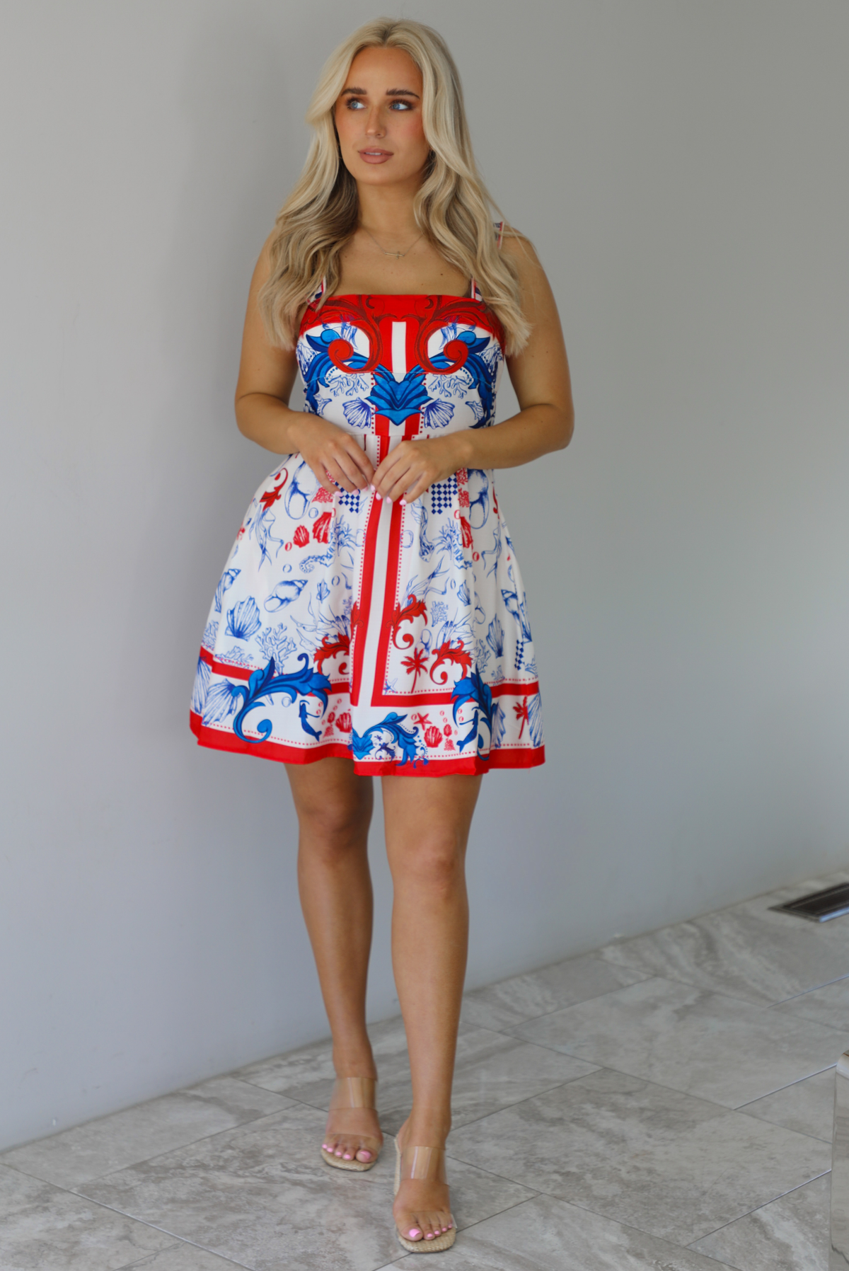 Woman wearing a white dress with red and blue sea themed graphics throughout with thick straps and functional pockets on either side. The photo is taken indoors against a white wall with white marble flooring. 
