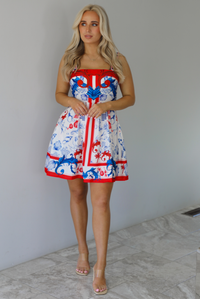 Woman wearing a white dress with red and blue sea themed graphics throughout with thick straps and functional pockets on either side. The photo is taken indoors against a white wall with white marble flooring. 