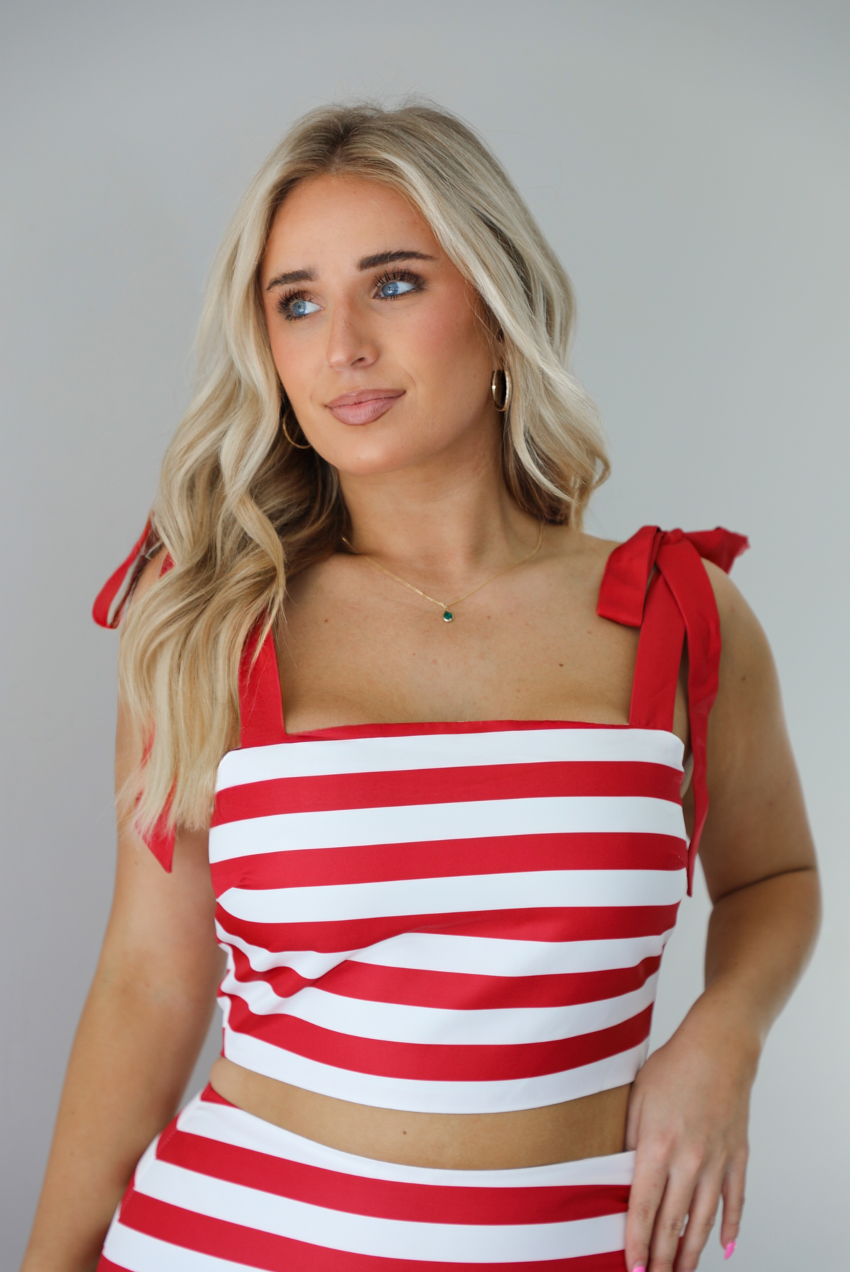 Woman wearing a red and white striped two piece set featuring a cropped top and maxi length, high waisted skirt. The photo is taken indoors against a white wall and shows a closeup of the top os the set, which is cropped and has red ribbon straps that are adjustable at the top of the shoulder. 
