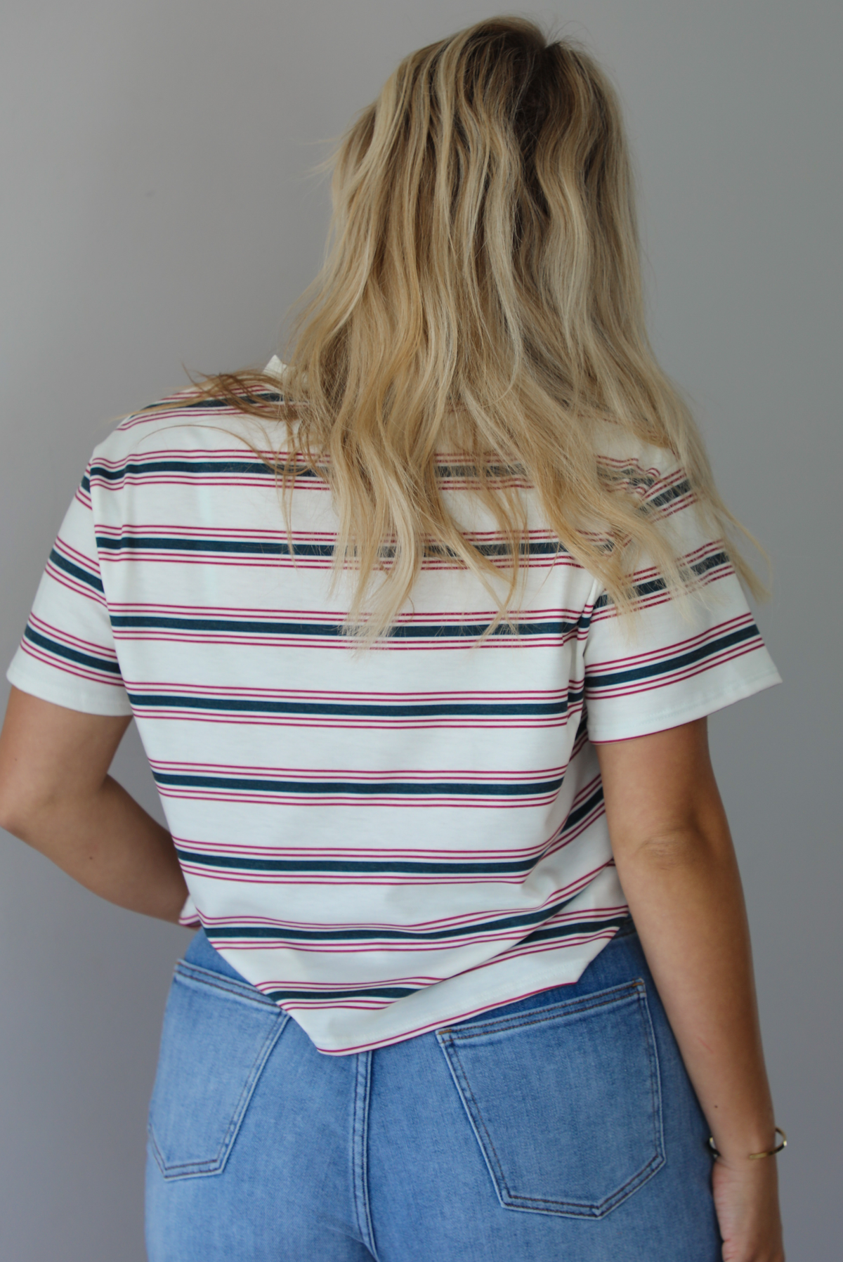 Woman wearing a striped, slightly cropped tee with light-wash denim shorts indoors against a white wall. The photo shows the back of the tee. 
