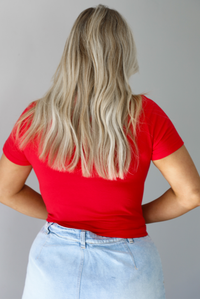 Woman wearing a red fitted baby tee with an embroidered horse graphic on the front. The top is paired with a high waisted, light-wash denim skort. The photo is taken indoors against a white wall and shows the back of the top. 