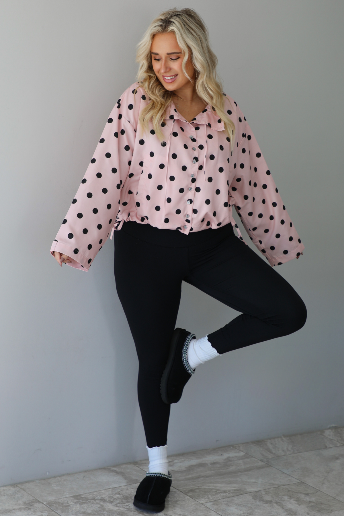 A woman wearing a pink button-down jacket with a black polka dot print pattern, paired with black leggings, white ankle socks, and black slide on slippers, standing indoors against a white wall with white marble flooring. 