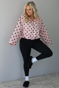 A woman wearing a pink button-down jacket with a black polka dot print pattern, paired with black leggings, white ankle socks, and black slide on slippers, standing indoors against a white wall with white marble flooring. 