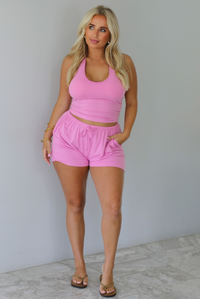 A woman wearing a matching pink activewear set consisting of a halter crop top and drawstring shorts, standing indoors against a white wall with white marble flooring.