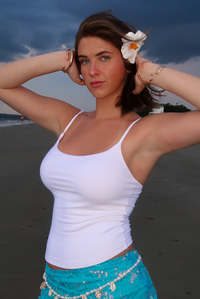 A woman posing on a beach wearing a white spaghetti-strap tank top and a turquoise belly dance skirt with gold coin accents, with a white flower in her hair.