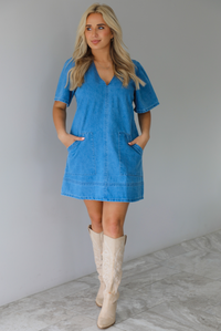 Woman wearing a v-neck, short sleeved, denim mini dress with functional pockets on either side. The photo is taken indoors against a white wall with white marble flooring. 