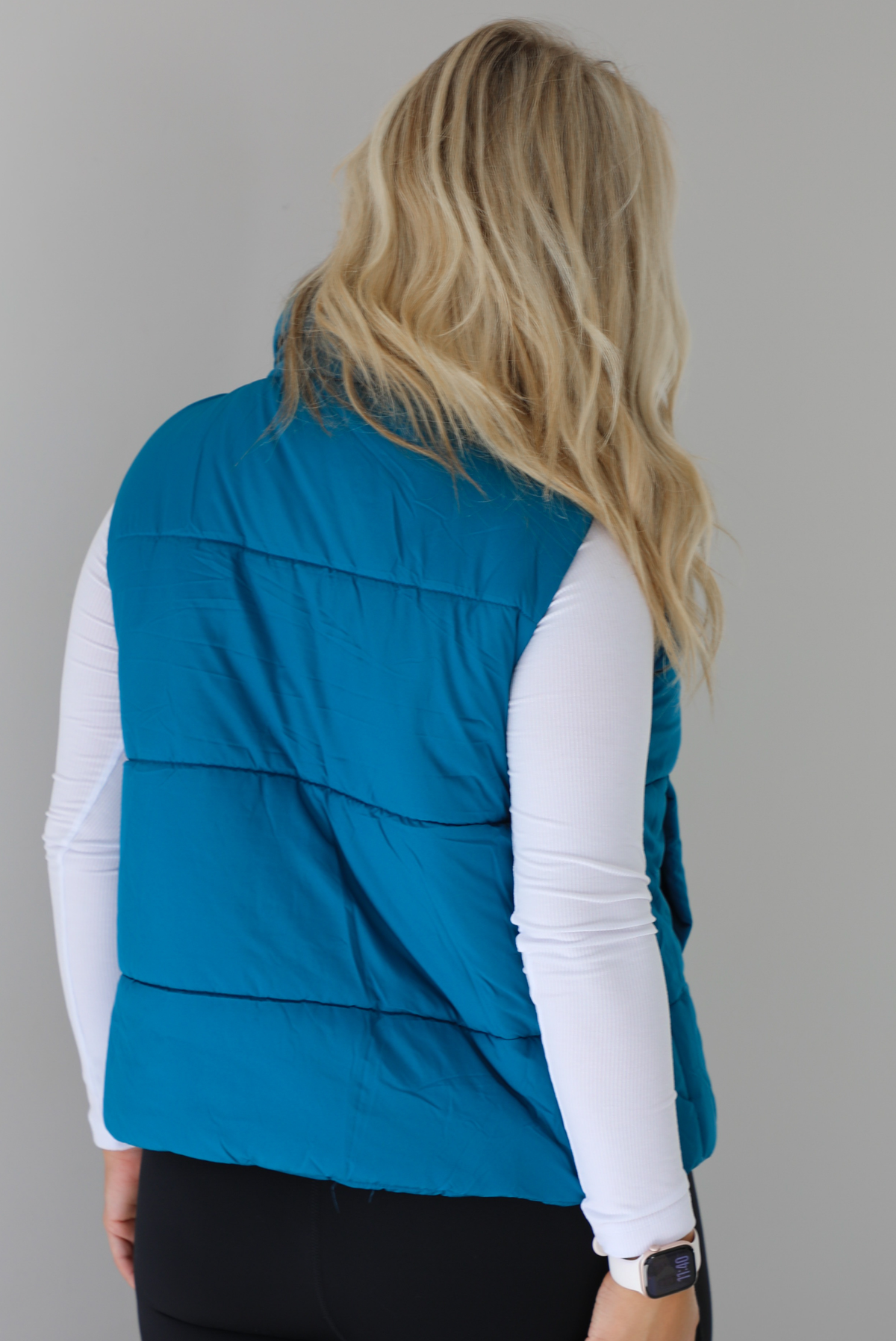 Woman wearing a teal puffer vest, white long-sleeve shirt, and black leggings indoors against a white wall. The puffer vest has a collared design, functional button closures at the neckline, an unzipped closure in the front, and functional pockets on either side. The photo shows the back of the puffer vest.