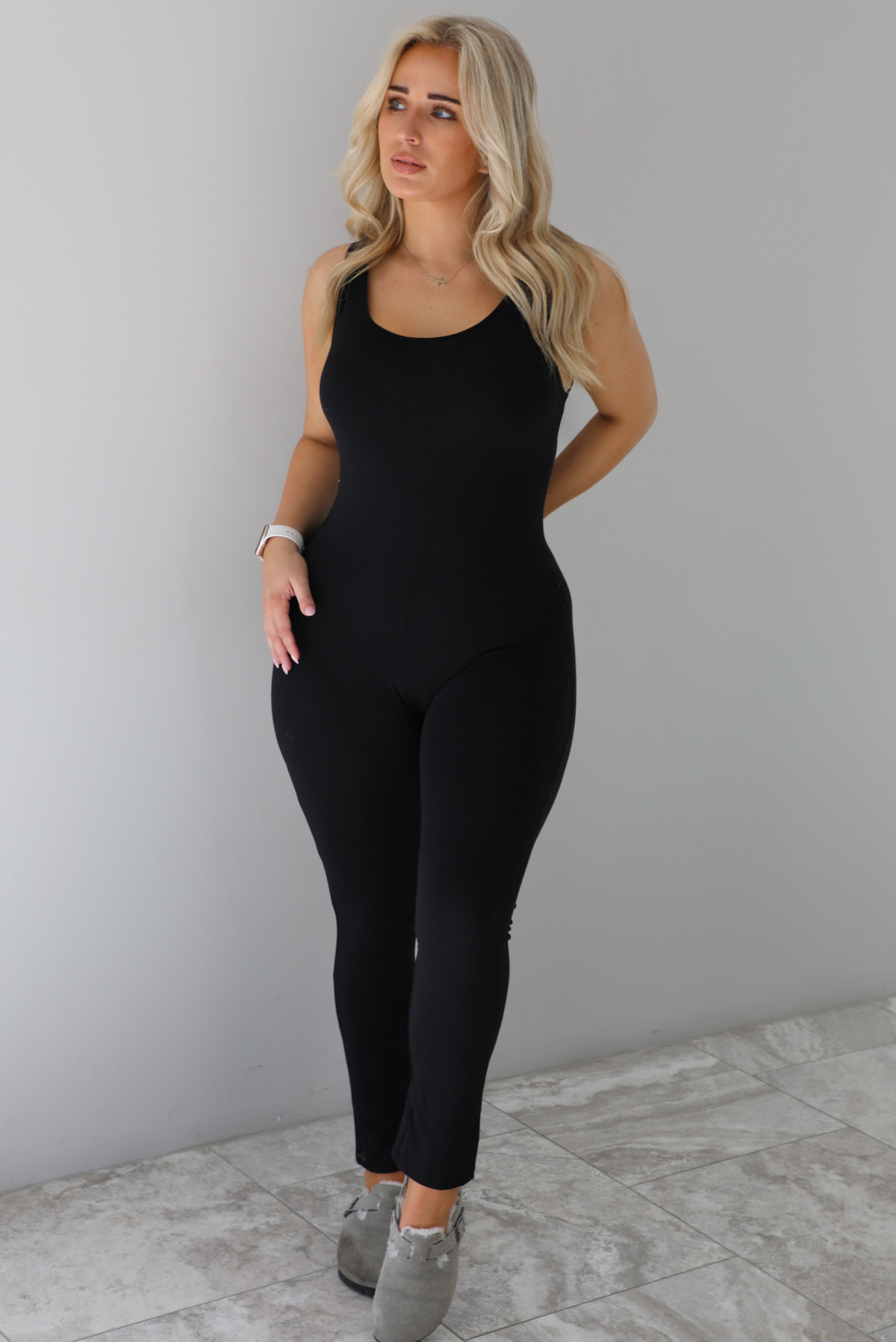 Woman wearing a black fitted jumpsuit with thick tank-top style straps. The jumpsuit is paired with grey slide on shoes. The photo is taken indoors in front of a white wall with white marble flooring. 