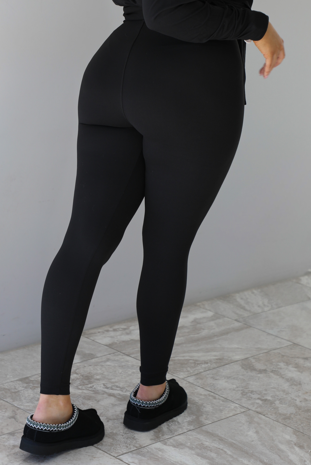 Woman wearing a black two piece set that features an oversized long sleeved top and fitted leggings. The photo shows a closeup of the back of the leggings. The photo is taken indoors against a white wall with white marble flooring. 