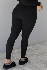 Woman wearing a black two piece set that features an oversized long sleeved top and fitted leggings. The photo shows a closeup of the back of the leggings. The photo is taken indoors against a white wall with white marble flooring. 