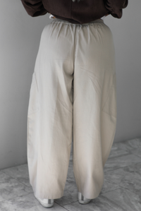 A woman shown from the waist down from the back wearing a pair of sand baggy pants, standing indoors against a white wall with white marble flooring. 