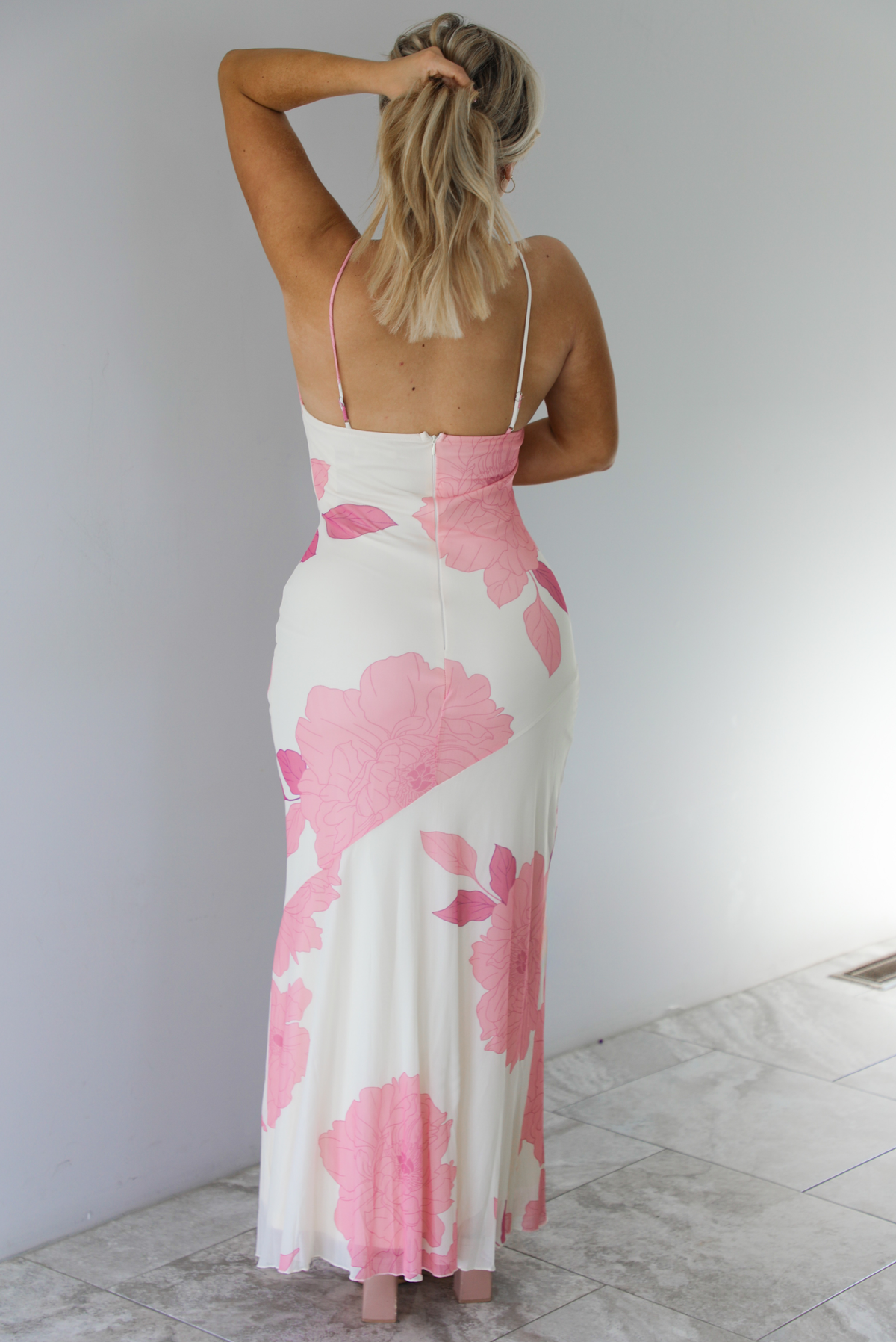 Woman wearing a cream maxi dress with a pink floral design throughout, and a halter neckline with a gold buckle detail. The photo is taken indoors against a white wall with white marble flooring and shows a closeup of the back of the dress with its adjustable straps and zipper closure. 