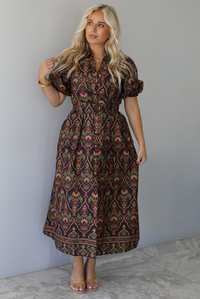 A woman wearing a dark brown midi dress with a multi-colored design throughout, a collared neckline, short puffed sleeves, functional button closures along the front, a belted detail at the waistline, standing indoors against a white wall with white marble flooring. 