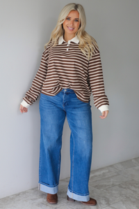 A woman wearing a brown and cream striped collared quarter-button top, paired with dark, wide leg denim with a cuffed hem, standing indoors against a white wall with white marble flooring. 
