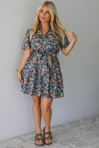 A woman modeling a short-sleeve floral mini dress with a tie waist, standing indoors against a white wall with white marble flooring. 