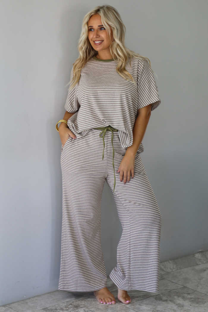 A woman modeling a matching two-piece loungewear set consisting of a short-sleeve top and wide-leg pants, both featuring thin beige and olive green horizontal stripes, with an olive green drawstring waist, standing indoors against a white wall with white marble flooring. 