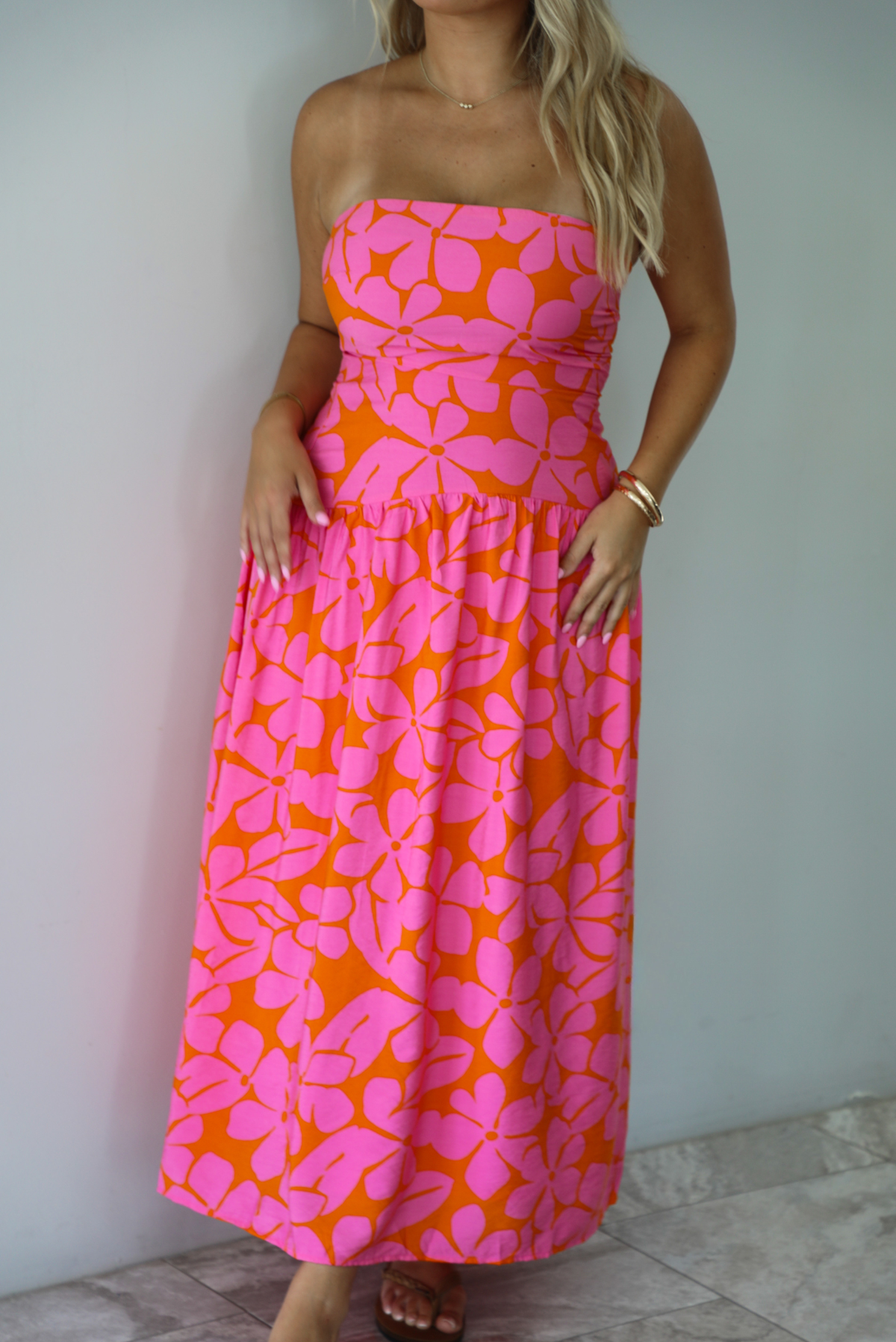 A woman modeling a strapless midi dress with a vibrant pink and orange floral pattern, standing indoors agains a white wall with white marble flooring. 