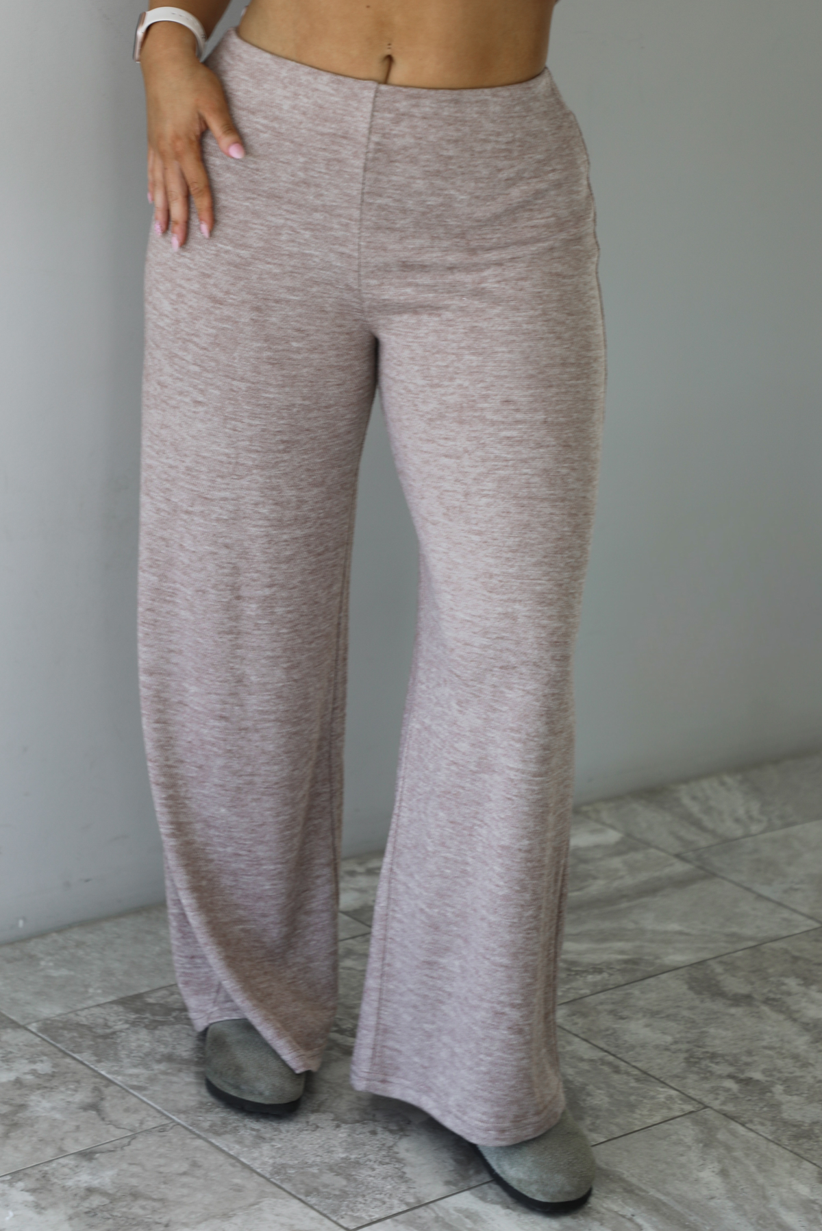 Woman wearing a taupe colored two piece set featuring a vest-appearance top with non-functional button closures on the left side and high waisted pants. The photo shows a closeup of the bottoms. The photo is taken indoors against a white wall with white marble flooring. 