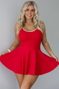 Woman wearing a red athleisure dress that has white trim at the top of the bodice, a fitted bodice, and flowy skirt. The photo is taken indoors against a white wall.